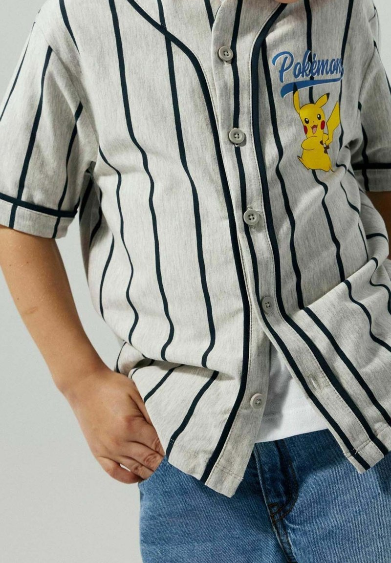 Gros plan d'une personne portant une chemise grise à rayures boutonnée avec un motif Pikachu et le mot Pokémon sur la poitrine, associée à un jean bleu.
