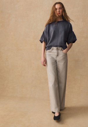 Fiatal nő hosszú hajjal, sötétszürke puffos ujjú felsőt, világos bézs széles szárú farmert és fekete cipőt visel, egy egyszínű háttér előtt áll.