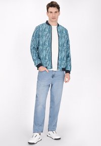Giacca bomber blu con un design a motivi, polsini neri a coste, abbinata a pantaloni in denim chiaro e sneakers bianche.