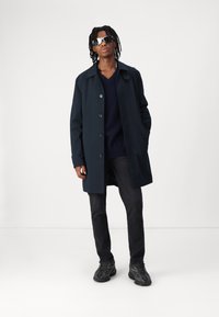 Marineblauwe waterdichte trenchcoat met een kraagontwerp, vijf knopen en zijzakken, gecombineerd met een donkere sweater en jeans.