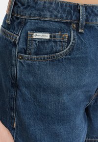 Shorts in denim blu scuro con leggere sfumature, dotati di una tasca posteriore con un'etichetta "Guess Jeans" visibile e cuciture arancioni a contrasto.