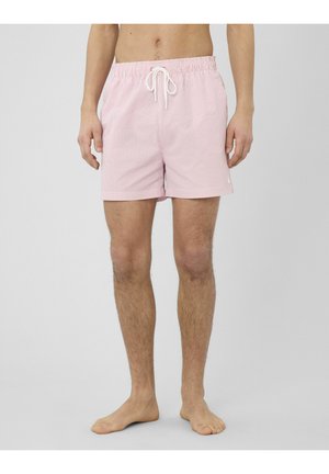 Homme portant un short de bain rayé rose et blanc avec une taille à cordon, debout pieds nus sur un fond gris uni.
