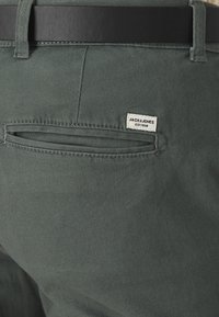 Ciemnozielone bawełniane spodnie o teksturowanym wykończeniu, z tylną kieszenią i białą metką z napisem "JACK & JONES EST. 1990." Widoczny czarny pasek.