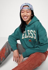 Felpa teal con testo bianco e giallo, abbinata a pantaloni tessuti arancio-marrone. Un cappellino multicolore poggia su capelli scuri.