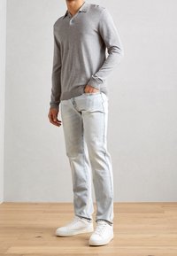 Polo shirt grigio a maniche lunghe con colletto e due bottoni, abbinato a jeans azzurri chiari e sneakers bianche, in piedi su un pavimento di legno.