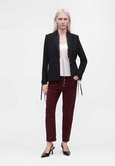Femme aux longs cheveux blancs portant un blazer noir avec bordure en dentelle, un haut blanc, un pantalon bordeaux avec des boutons devant et des talons noirs, debout sur un fond blanc.