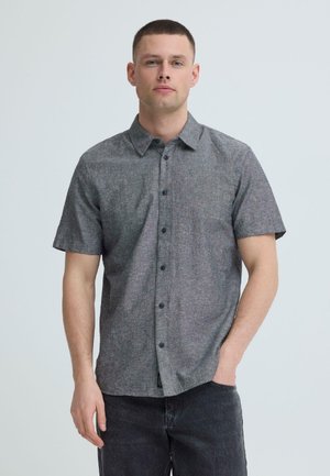 Uomo con camicia grigia a maniche corte con bottoni e jeans scuri, in piedi con una mano in tasca contro uno sfondo semplice.