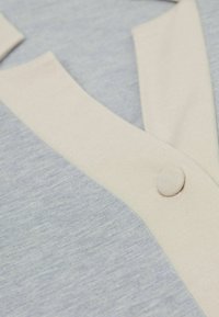 Ljusblått tyg med en strukturerad väv, som har en krämfärgad krage och knapp. Designen inkluderar överlappande tygstycken.