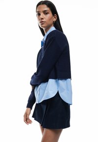 Meriino villaneule tummansinisellä pohjalla, jonka päällä vaaleansininen paita pyöreällä helmalla, yhdistettynä tummansinjaan hameeseen. Neuleessa on teksturoitu yksityiskohta.