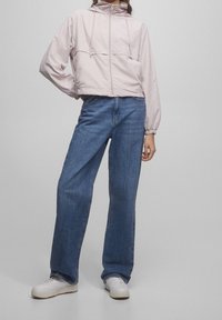 Light pink cropped jacket with drawstrings at the collar, paired with wide-leg blue jeans and white sneakers, set against a neutral background.