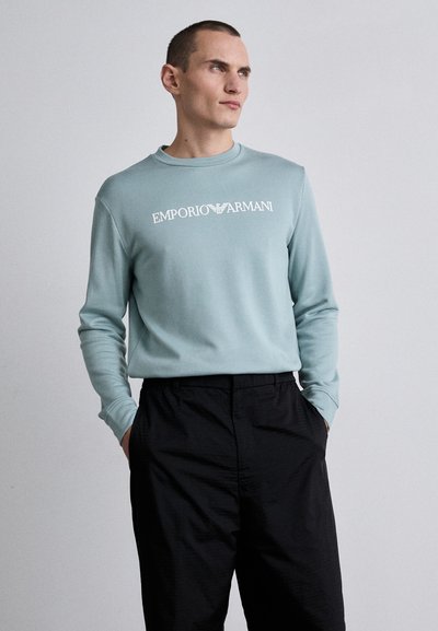 Hele sinine kapuutsita sviiter pikkade varrukatega ja ümarate kaelustega, millel on rinnal valge "Emporio Armani" logo. Mustad püksid.