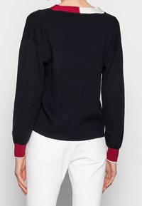Person in einem schwarzen Pullover mit rotem und weißem Kragen sowie Bündchen, kombiniert mit weißen Hosen, von hinten vor einem schlichten Hintergrund gezeigt.