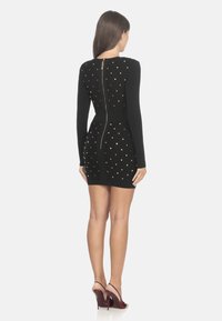 Abito nero aderente con maniche lunghe, caratterizzato da borchie dorate disposte in un motivo a diamante e una zip centrale sul retro. Indossato con sandali con tacco color mogano.