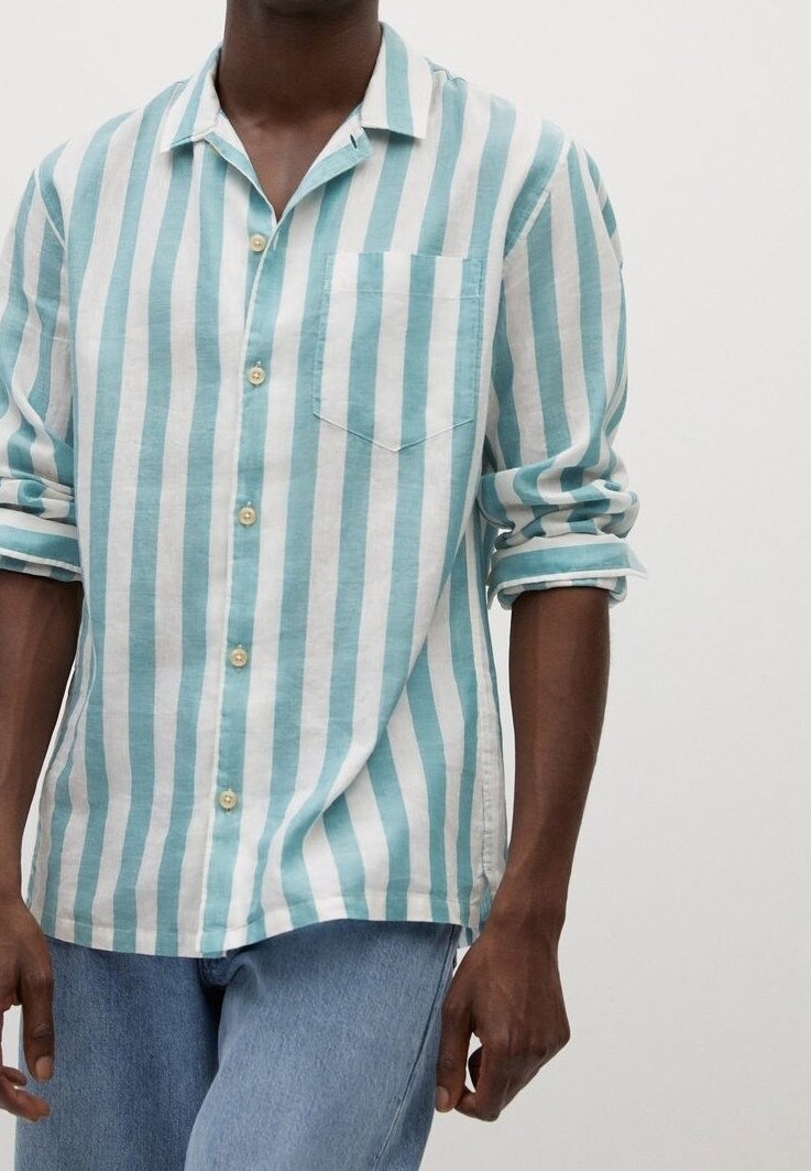 Homme portant une chemise à boutons rayée verticalement bleu clair et blanc avec les manches retroussées, et un jean bleu clair, devant un fond uni.