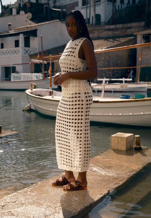 Žena s dugom pletenicom, koja nosi bijelu kroše haljinu bez rukava i sandale s uzorkom, stoji na pristaništu pokraj čamaca i vode.