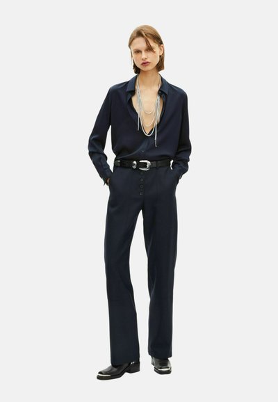 Frau trägt ein marineblaues Hemd mit Knöpfen und geschichteten silbernen Halsketten, hochgeschnittene marineblaue Hose mit Knöpfen, schwarzen Gürtel und schwarze Stiefel.