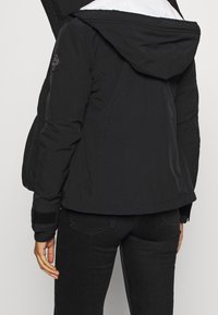 Svart vadderad jacka med huva, med en diamantpatch på vänster arm och ribbade mudd. Synlig vit foder vid kragen.