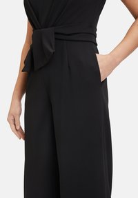Black jumpsuit featuring a wrap design with a knotted accent at the waist and pockets. Made from smooth, lightweight fabric.