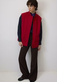 Žena stojí proti bielej stene a má na sebe červenú bezrukávovú vestu cez čiernu košeľu s dlhým rukávom, hnedé široké nohavice a čierne topánky.