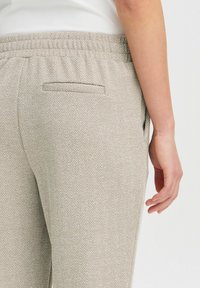 Lys beige bukser med fiskebensmønster, elastisk talje og en enkelt baglomme, vist sammen med en hånd.