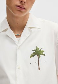 Witte shirt met korte mouwen met een groene palmbomenafbeelding en daaronder de tekst "HUGO". De stof lijkt lichtgewicht en glad te zijn.