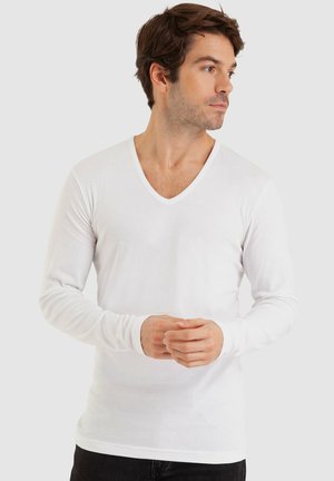 Chemise blanche à manches longues en V, fabriquée dans un matériau doux, dotée d'un design ajusté et de poignets côtelés, présentée sur un mannequin avec un fond neutre.