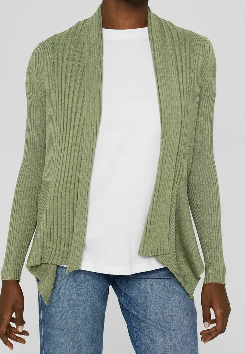 Personne portant un cardigan vert clair côtelé ouvert sur un chemisier blanc et un jean bleu, debout devant un fond uni.