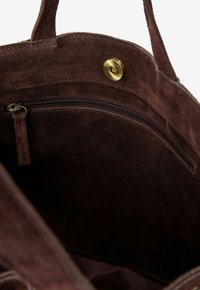 Bruine suède interieur van een tas met een zichtbare ritssluiting en een ronde messing drukknoop. Zachte textuur en rijke kleurstelling zijn opvallend.