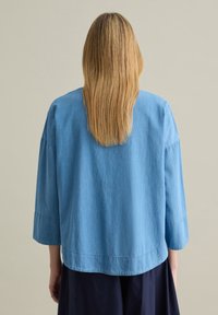 Camicia azzurro chiaro, dalla vestibilità ampia, a maniche 3/4 con scollatura rotonda e texture sottile. L'orlo è dritto con una leggera discesa sul retro.