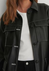 Chaqueta de piel sintética negra con costuras blancas, que cuenta con dos bolsillos frontales, un cierre con botones y un diseño con cuello, sobre una camiseta blanca de canalé.