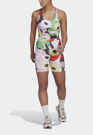 Kvinde iført farverig, blomster-mønstret sportstop og shorts, hvide sokker og blomstrede sneakers, står mod en ensfarvet baggrund.