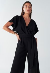 Czarny kombinezon kopertowy z krótkimi, falbanowymi rękawami, głębokim dekoltem w serek i dopasowanym paskiem w talii. Gładka tkanina z szerokimi nogawkami.