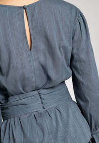 Vestido azul a rayas con un cierre en la parte posterior en forma de ojal, mangas fruncidas y una cintura ceñida con cinturón y detalles de botones. Tejido suave y texturizado.
