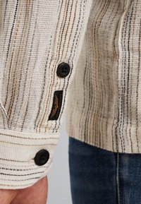 Chemise en tissu tissé avec un tissu texturé crème, ornée de rayures verticales noires et brunes, dotée de boutons noirs et d'une étiquette de marque.