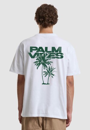 Osoba s kučeravými vlasmi, ktorá má na sebe biele tričko so zelenými grafikami palmy a textom "PALM VIBES" na chrbte, viditeľné béžové kraťasy.