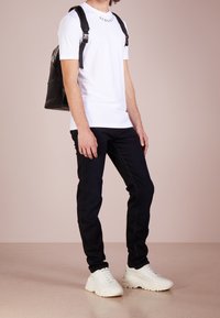 T-shirt bianco a maniche corte con logo "VERSACE", pantaloni in denim neri, zaino nero e sneakers bianche chunky. Tessuto morbido e design attillato.
