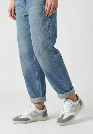 Osoba oblečená v ohrnutých modrých džínách a bílých teniskách se šedými semišovými detaily, stojící s jednou nohou mírně zvednutou.