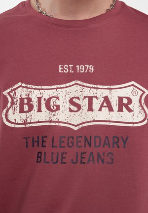 Raudona medvilninė marškinė su nusidėvėjusio logotipo dizainu kreminėmis ir tamsiai mėlynomis spalvomis, kuriame užrašyta "BIG STAR EST. 1979 LEGENDINIAI MĖLYNI DŽINSAI."