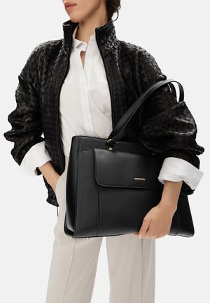 Donna che tiene una grande borsa tote in pelle nera, indossa una camicia bianca e una giacca in pelle intrecciata nera con le maniche arrotolate.