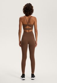 Rjav športni crop top z zasnovo zvitega hrbta, v kombinaciji z visokimi rjavimi pajkicami. Vključuje črne superge. Gladka tekstura.