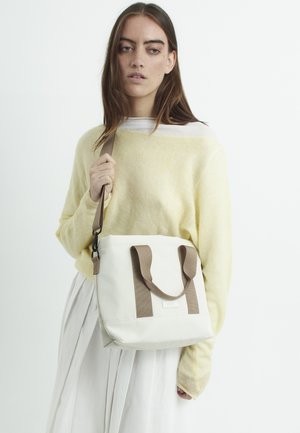 Weißes Tote Bag mit beigen Trägern und strukturierten Details. Das Model trägt einen gelben Pullover und einen weißen Rock und hält die Tasche.