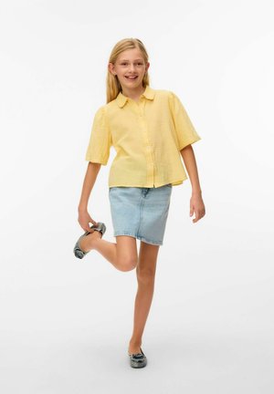 Fiatal lány sárga blúzban és farmer szoknyában, cipőt tartva a felemelt lába mögött, mosolyog egy egyszerű fehér háttér előtt.