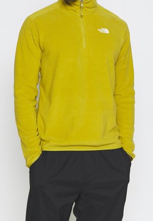 Personne portant une polaire jaune moutarde à fermeture éclair quart, avec le logo The North Face sur la poitrine, pantalon noir, mains dans les poches, debout devant un fond blanc.