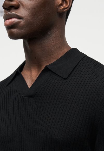 Polo en maille côtelée noire avec col en V et col plat, doté d'une coupe ajustée qui met en valeur la texture et la forme.