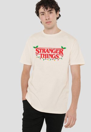 Junger Mann, der ein beiges "Stranger Things"-T-Shirt trägt, das mit Weihnachtslichtern und Stechpalmenblättern verziert ist, steht mit einer Hand in der Tasche.