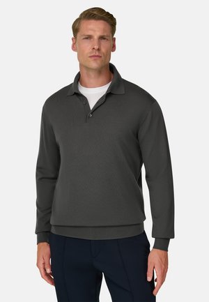 Homme blond portant un polo gris foncé à manches longues sur un maillot blanc et un pantalon sombre, debout devant un fond uni.