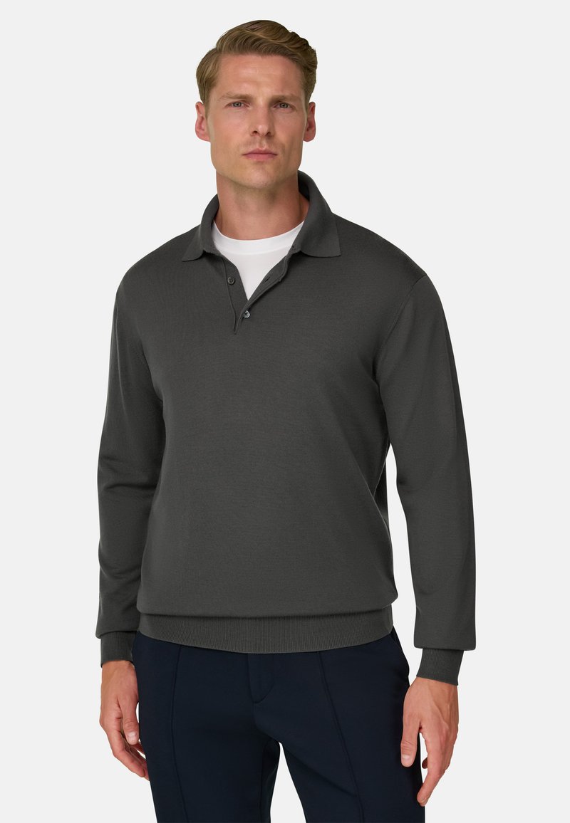 Homme blond portant un polo gris foncé à manches longues sur un maillot blanc et un pantalon sombre, debout devant un fond uni.