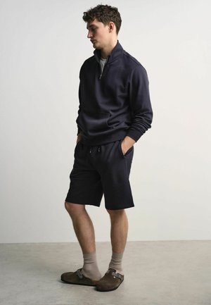 Man wearing navy quarter-zip sweatshirt, navy shorts, beige socks, and brown slip-on sandals standing with hands in pockets.