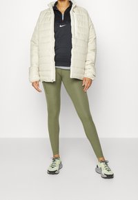 Puffer-takki vaaleassa, musta pitkähiha, oliivinvihreät legginsit ja harmaat lenkkarit neonkorostuksilla. Teksturoitu materiaali; urheilullinen istuvuus.