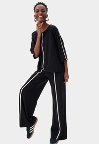Zwarte loszittende top en wijde broek met witte zijstrepen. Het materiaal lijkt zacht. Model draagt donkere sneakers met lichte zolen.